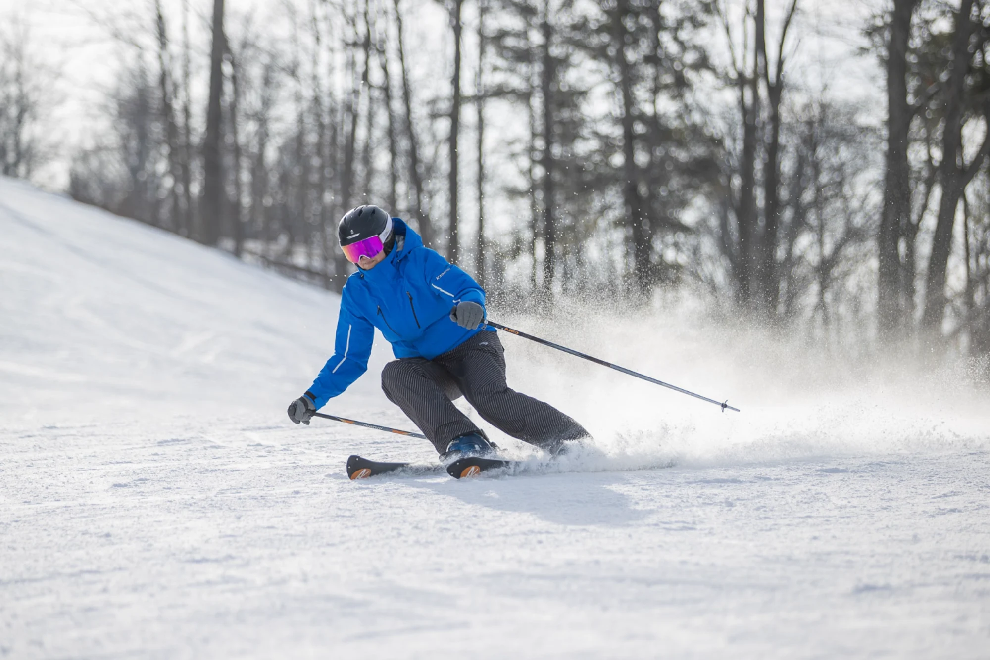 A skier wearing a black helmet and pink goggles, dressed in a blue jacket and dark grey snowpants, making a turn on a snow-covered run with trees in the background